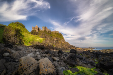 Dunluce Castle seen from behind of mossy rocks on a shore, nested on the edge of cliff, part of...