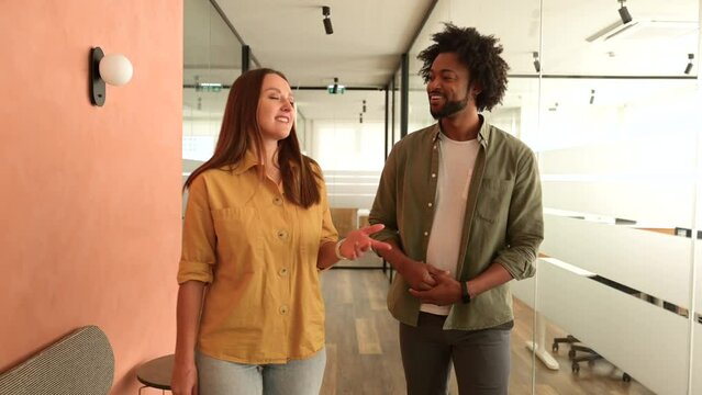 Young enthusiastic female colleague explains new strategy to male employee and gesturing. Indoor portrait of diverse work team discussing, walking through hallway in modern coworking office space