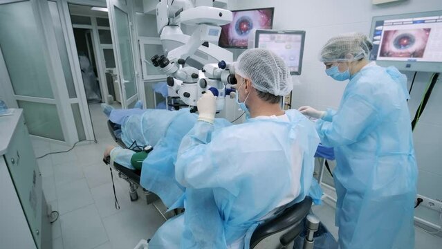 Ophthalmological surgery specialist sits in front of microscope performing operation. Low angle view on the female doctor conducting high-precision surgery. Femto Laser Assisted Cataract Surgery.