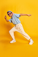 Fototapeta na wymiar Dynamic portrait of little african boy, hip-hop dancer in stylish street style clothes dancing over bright yellow background. Concept of music, dance happiness