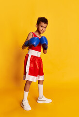Active sportive little boy, beginner boxer in sports uniform and boxing gloves isolated over yellow...