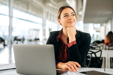 Positive caucasian businesswoman, office employee, product manager, sits in a business center, working in a laptop on a new project, looking away, dreaming about vacation, smiling