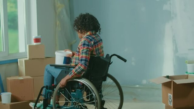 A young African American woman in a wheelchair is talking on a mobile phone. The disabled woman takes a bucket of paint and a roller and sharing plans to repairs a room. Handicapped person.