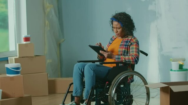 Young African American woman in a wheelchair is planning a renovation using a digital tablet. Disabled woman browses interior design ideas on the internet. Handicapped person.