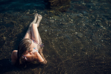 A blonde girl with long hair, in a striped swimsuit, by the ocean, sits by the rocks, in the water, on a bright sunny day.