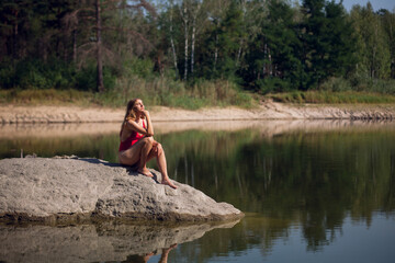 A young girl with long blond hair, with a slender figure, in a red bathing suit, sits on a stone, in a river, against the backdrop of a forest.