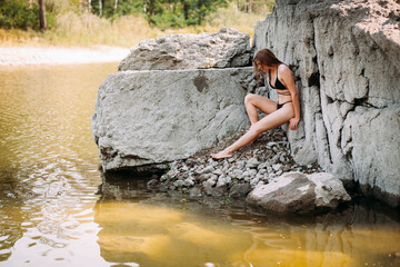 A beautiful girl with long blond hair, a slender figure, in a black swimsuit, sits on the rocks near the ocean on a hot sunny summer day.