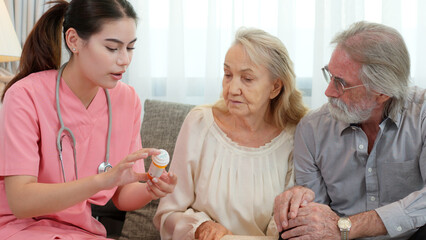 Caucasian young adult nurse is visiting elderly couple and holding a medicine bottle to explain how...