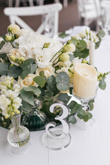 Composition for a festive wedding tables of white flowers of hydrangea, rose, eustoma and eucalyptus, different candles and a table number. Wedding outdoor concept