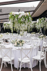 A general plan of a festive tables decorated with a white tablecloth, white chairs, plates, compositions of white flowers and candles. Hanging flower beds and light bulbs. Wedding decoration and decor