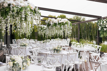 A general plan of a festive tables decorated with a white tablecloth, white chairs, plates, compositions of white flowers and candles. Hanging flower beds and light bulbs. Wedding decoration and decor