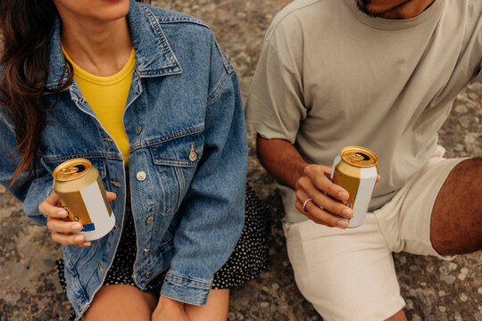 Midsection of male and female friends holding drink cans while sitting together on rock