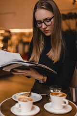 Concentrated young woman in black long sleeves top and eye glasses with coffee cup reading magazine in the coffee shop. Woman with two cup of coffee, glass water and cake waiting for someone.