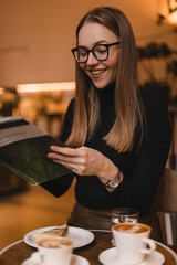 Happy young woman in black long sleeves top and eye glasses reading magazine in the coffee shop. Woman with two cup of coffee, glass water and cake laughing while see something in journal.