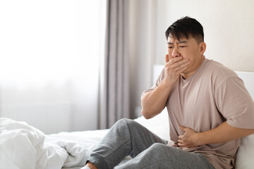 Sick korean man sitting in bed, covering mouth, touching belly