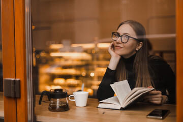 Woman wear black sweater and eye glasses, reading a paper book with a cup of tea in outdoor cafe,...