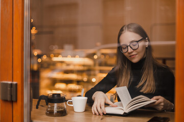 Woman wear black sweater and eye glasses, reading a paper book with a cup of tea in outdoor cafe,...