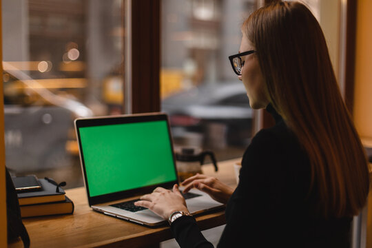 Side view blonde woman freelancer in eyeglasses working in cafe, using laptop computer with a green screen on monitor. Concept remote work, freelance, working on laptop computer or net-book.