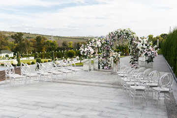 Wedding ceremony outdoor. A beautiful and stylish wedding arch, decorated by various fresh white flowers with transparent chairs, standing in the garden. Celebration day.