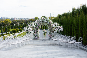 Wedding ceremony outdoor. A beautiful and stylish wedding arch, decorated by various fresh white flowers with transparent chairs, standing in the garden. Celebration day.