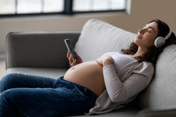 Obraz premium Calm Pregnant Lady In Wireless Headphones Listening Music On Smartphone At Home