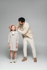 full length of bearded father adjusting crown of daughter in dress on grey.