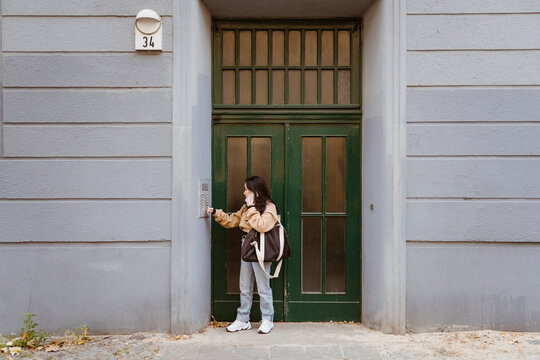 Woman talking on smart phone while doing intercom standing at apartment entrance