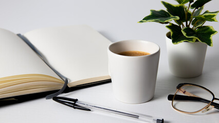Taza de café,  cuaderno blanco, gafas y bolígrafo sobre una mesa de madera blanca 