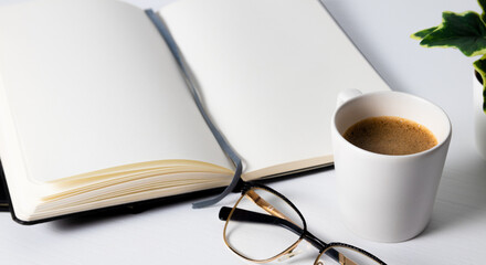 Taza de café,  cuaderno blanco, gafas y bolígrafo sobre una mesa de madera blanca 