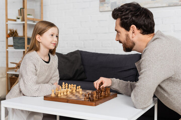 preteen girl smiling while playing chess with bearded dad at home.