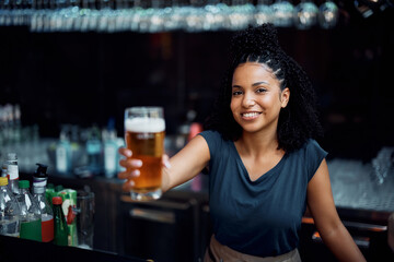 Happy African American waitress serving beer at bar counter and looking at camera.