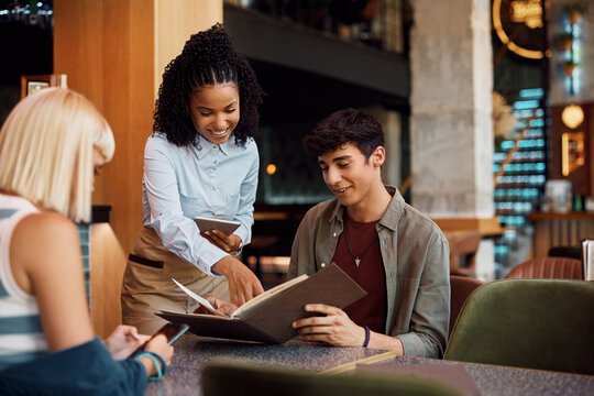 Happy black waitress recommends order from menu to her customers in cafe.