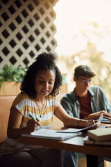 Young African American woman takes notes while studying with friend in cafe.