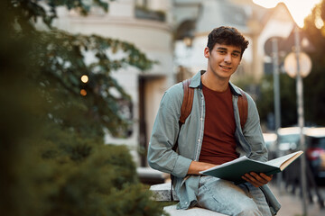 Smiling college student reading book at campus and looking at camera.
