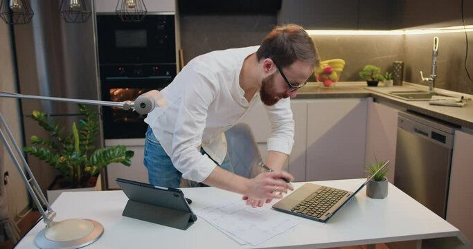 Man realtor in glasses draws up documents for a new property with a laptop stay at a table, making notes. Designers male are designing architectural structures for clients at home.