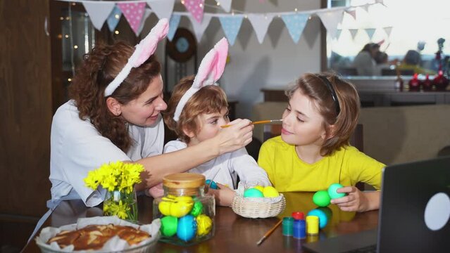 Easter Family traditions. Mom and two caucasian happy children with bunny ears dye and decorate eggs with paints for holidays while sitting together at home Easter Sunday table using laptop remote