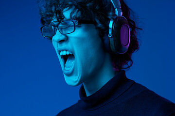 Teenage man wearing earphones listening to music and dancing and singing with glasses, hipster lifestyle, portrait blue background, neon light, style and trends, mixed light, copying space