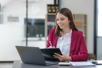 Happy businesswoman taking notes with laptop and document in office.