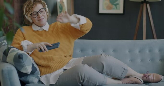 Portrait of carefree woman wearing headphones dancing having fun holding smartphone indoors at home. Modern technology and lifestyle concept.