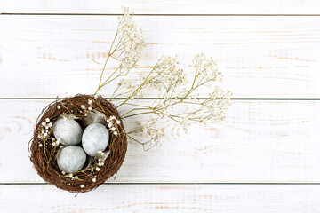 A brown nest of twigs with three gray Easter eggs, feathers and sprigs of gypsophila on a white wooden table. Minimalistic Easter concept