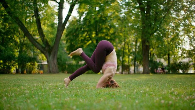 Active Woman does Headstand Yoga practice in Park. Fitness Lady exercises Sequence with Eagle pose outdoors among sunshine. Focus, Healthy Lifestyle and Balance. 4K wide orbit shot