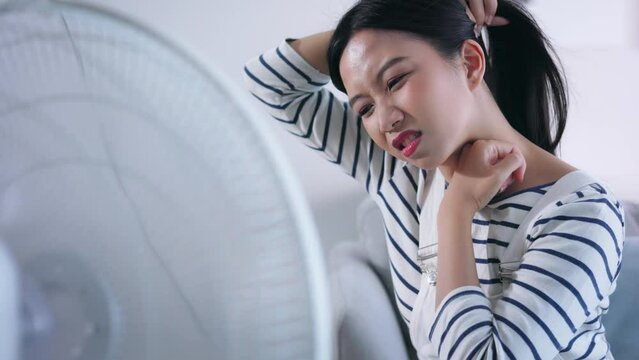 Young unhappy Asian woman suffering the summer heat and getting air flow from fan on sofa in living room at home
