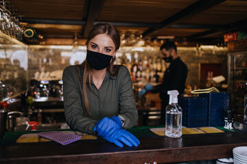 Portrait of a serious waitress standing behind a bar in a nice restaurant. She wears a protective mask and gloves as part of security measures against the Coronavirus pandemic.