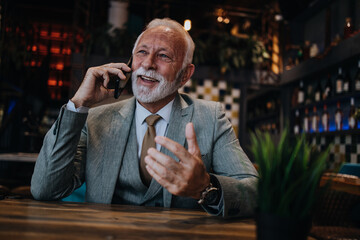 Happy businessman sitting in restaurant and waiting for lunch. He is using smart phone and talking with someone. Business seniors lifestyle concept.