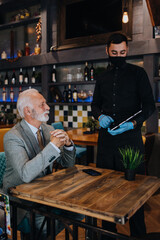 The waiter taking order for a delicious meal to the senior businessman at the restaurant. He wears a protective mask as part of security measures against the Coronavirus pandemic.
