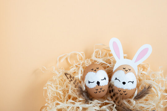 Sweet owl family. Brown easter eggs with painted faces and bird feathers. One owl in banny ears hairband. Beige background, place for your text.