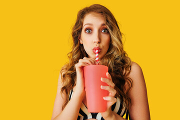 young adult woman holding pink glass and drinking straw over yellow background