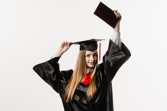 Happy girl student in black graduation gown and cap raises masters degree diploma above head on white background. Graduate girl is graduating college and celebrating academic achievement.