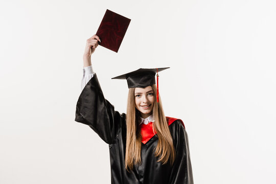 Graduate girl is graduating college and celebrating academic achievement. Happy girl student in black graduation gown and cap raises masters degree diploma above head on white background.