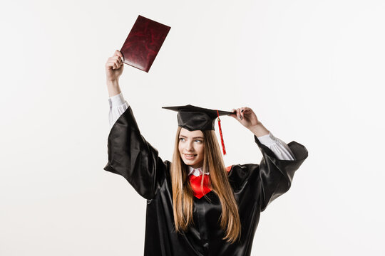 Graduate girl is graduating college and celebrating academic achievement. Happy girl student in black graduation gown and cap raises masters degree diploma above head on white background.